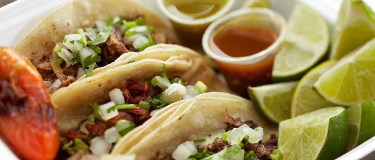 Mexican tacos in a container with limes and tomatoes.