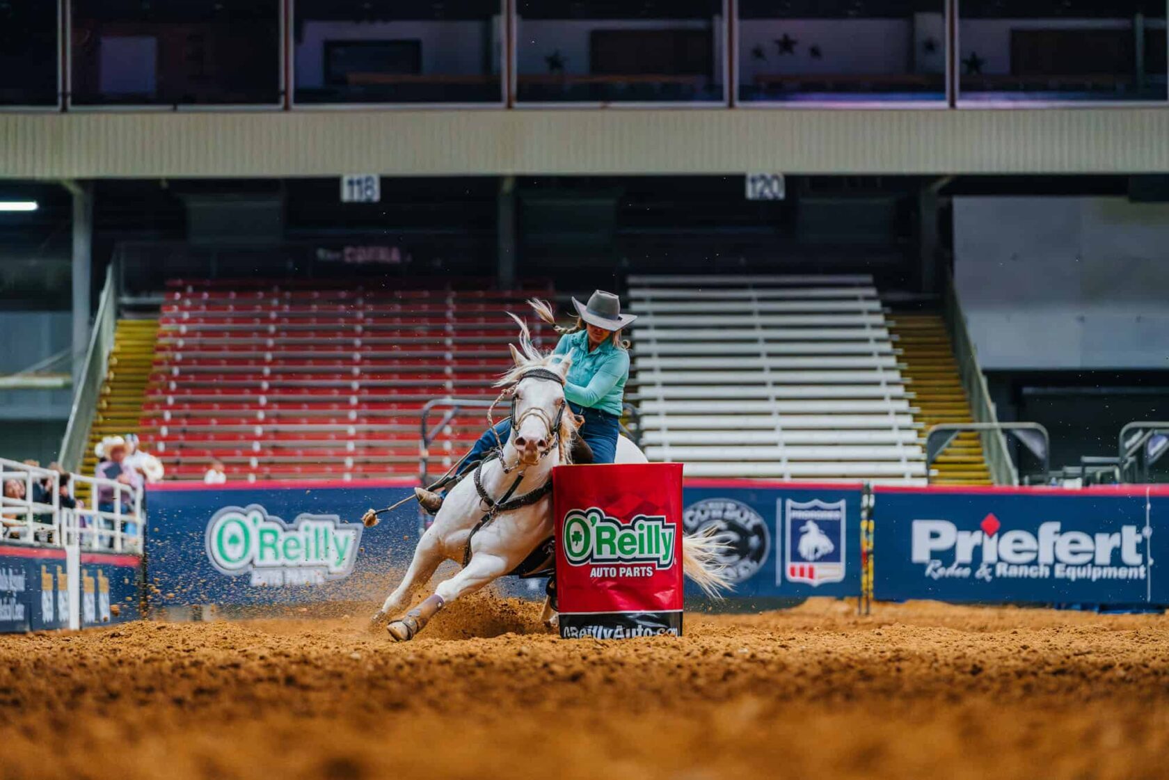 A rodeo competitor in a teal shirt and cowboy hat skillfully guides a galloping white horse around a red barrel in a dirt arena, showcasing a barrel racing event at a rodeo competition.