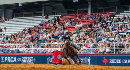 A rider on a horse competes in a barrel racing event at a rodeo in front of a crowded audience.