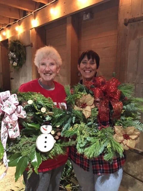 Two smiling women holding festive Christmas wreaths with red bows and a snowman decoration indoors with string lights above.