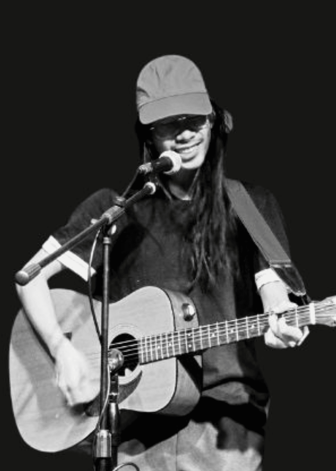 Person with long hair and a cap playing an acoustic guitar and singing into a microphone.