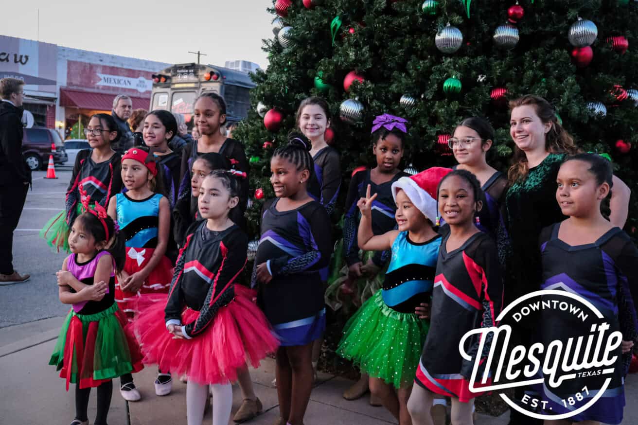 A group of children and adults in colorful outfits pose for a photo in front of a large decorated Christmas tree in Downtown Mesquite, Texas. A "Downtown Mesquite" sign appears in the corner.