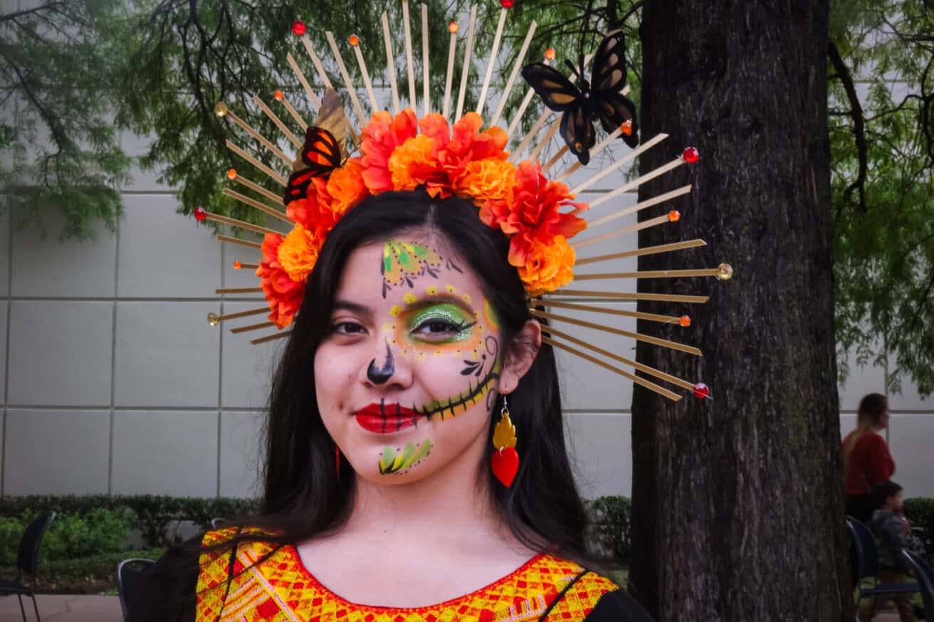 A woman wears vibrant makeup with skeletal designs and a headpiece adorned with flowers and butterflies, standing outdoors near a tree.