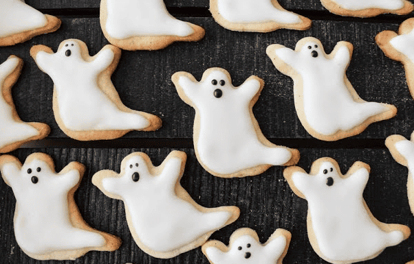 A batch of ghost-shaped sugar cookies with white icing and black eyes and mouths are arranged on a dark surface.