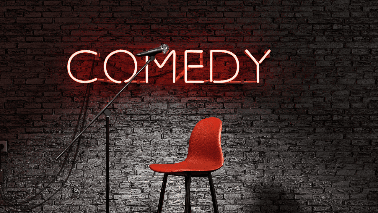 A red chair sits in front of a stand-up microphone, against a brick wall with a neon sign reading "COMEDY.