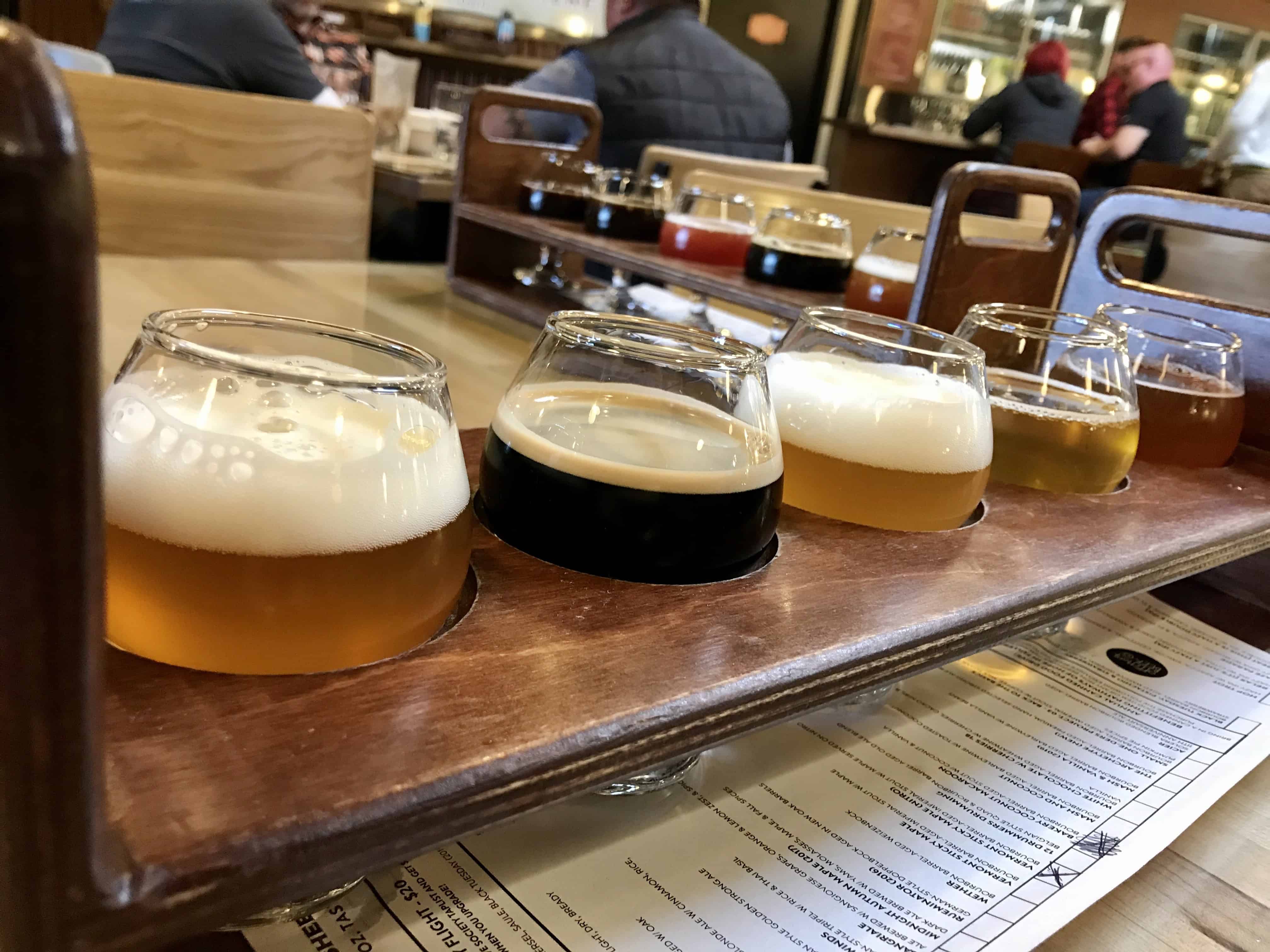 A wooden tray holds five glasses of beer in a restaurant setting. Each glass contains a different type and color of beer. Menu visible underneath tray.