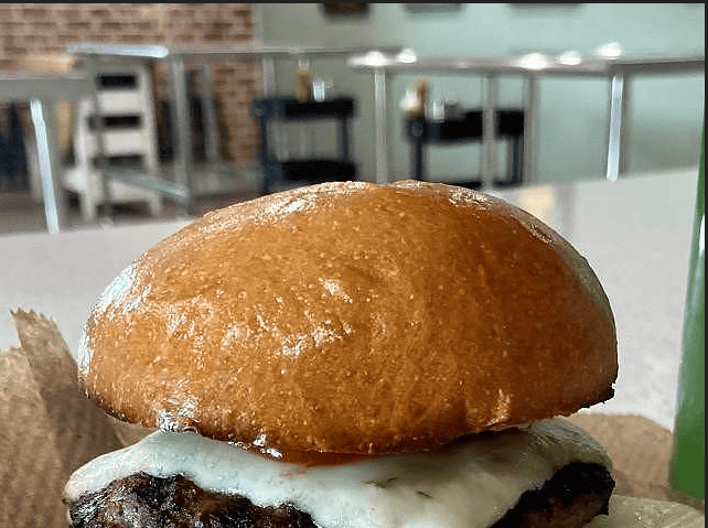 A close-up of a cheeseburger with a glossy bun on a table in a cafe setting. Tables, stools, and condiment bottles are visible in the background.