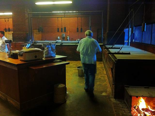 A worker in a white coat walks through a dimly lit brick smokehouse with large metal ovens and a fire burning in a hearth.