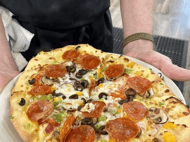 Person holding a plate with a freshly baked pizza topped with pepperoni, black olives, green peppers, mushrooms, and onions.