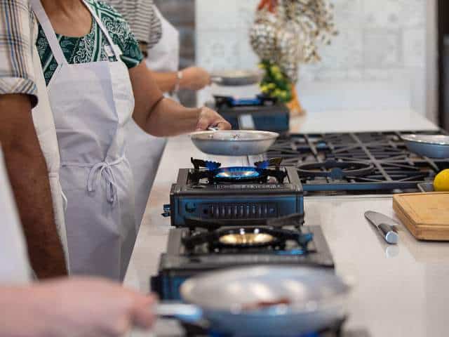 People in aprons cooking on portable gas stoves with frying pans in a kitchen setting.