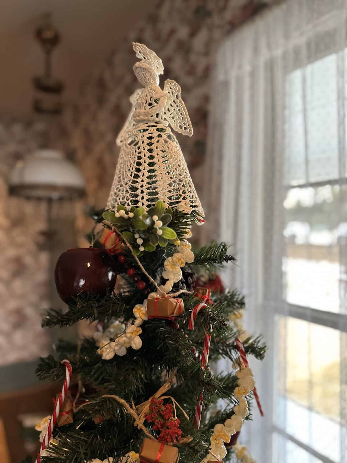 A Christmas tree topped with a crochet angel is decorated with apples, wrapped gifts, berries, and candy canes, next to a window.