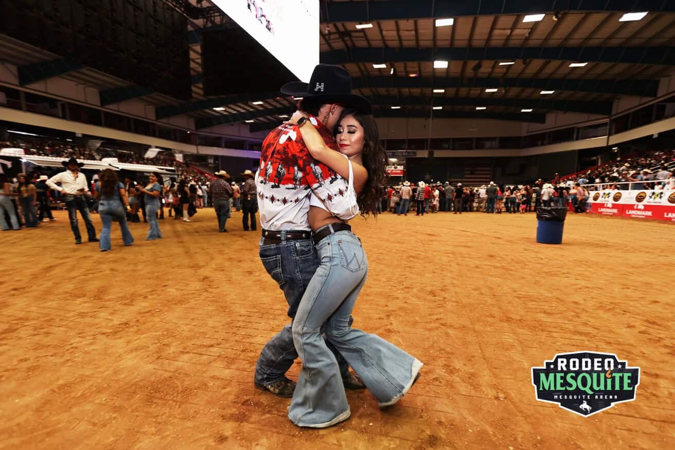 A couple in Western attire dances closely on a dirt floor inside an indoor arena, surrounded by spectators. A "Rodeo Mesquite" sign is visible in the corner.