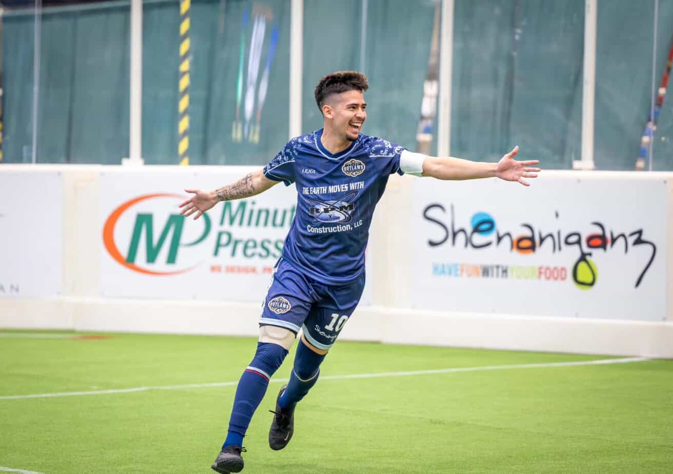 Soccer player in a blue uniform celebrates on the field with arms outstretched. Advertising boards visible in the background.