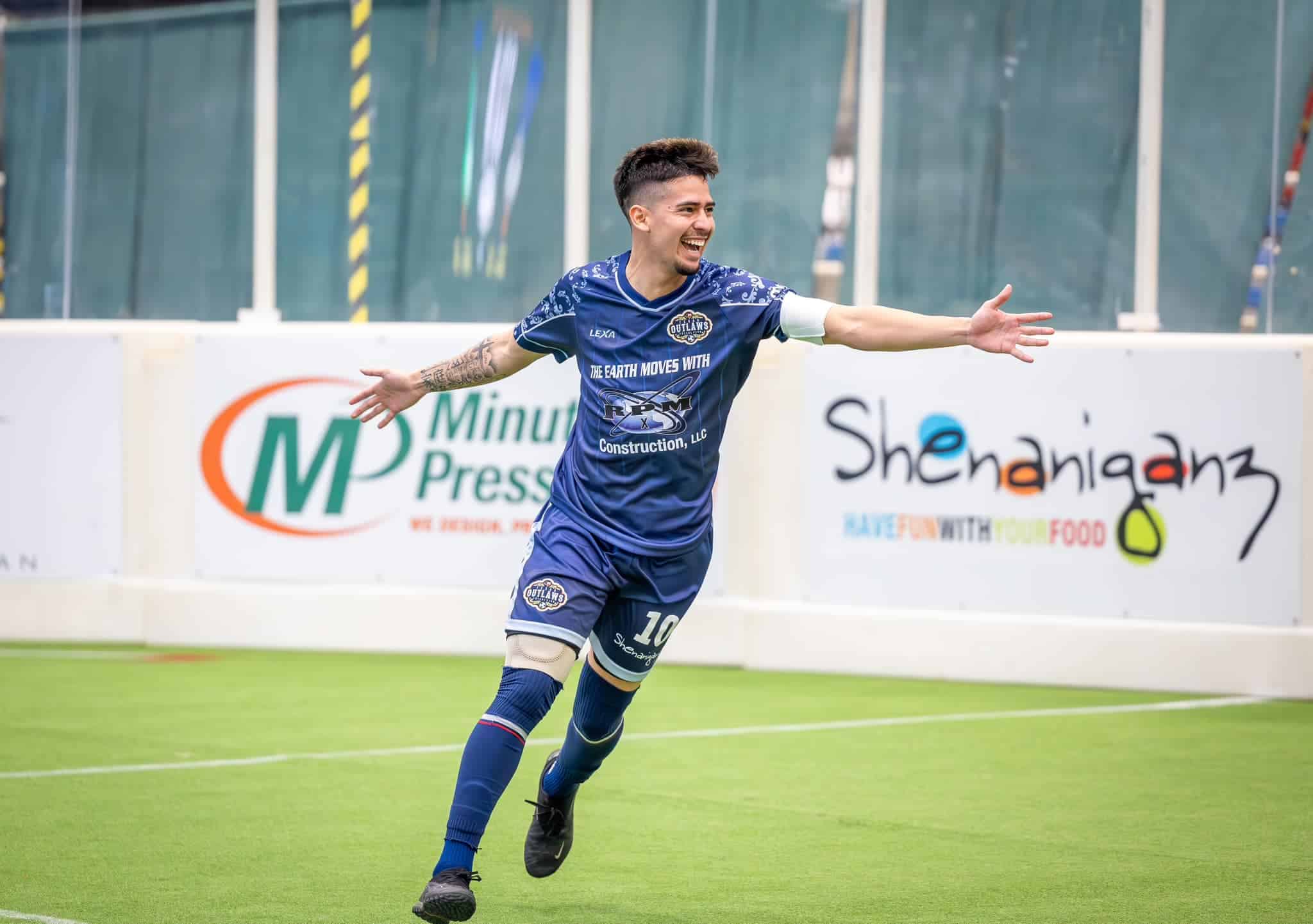 Soccer player in a blue uniform celebrates on the field with arms outstretched. Advertising boards visible in the background.