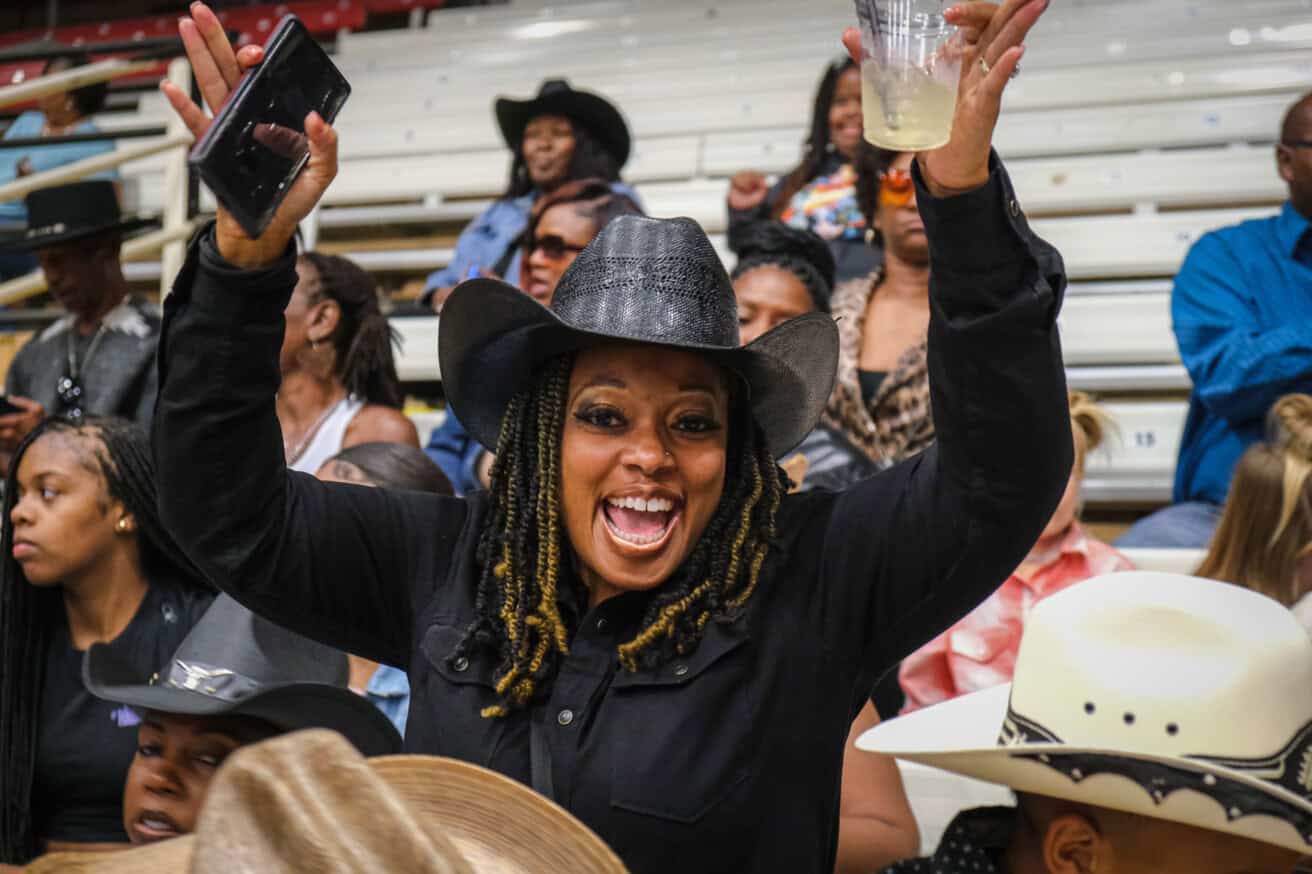 A woman in a black cowboy hat raises her arms, holding a phone and a drink, surrounded by a crowd on bleachers.