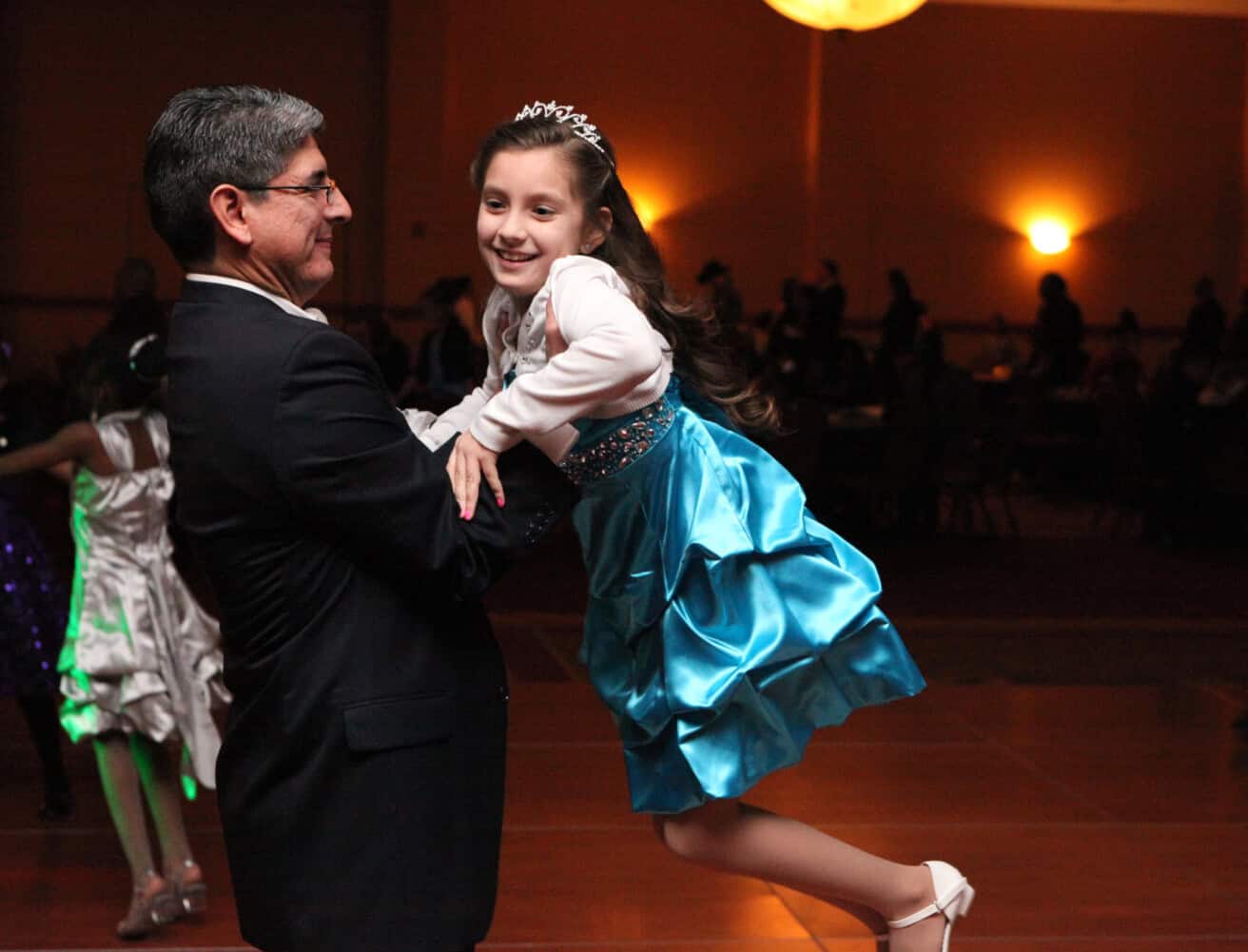 A man in a suit dances with a young girl in a blue dress and tiara on a dance floor.