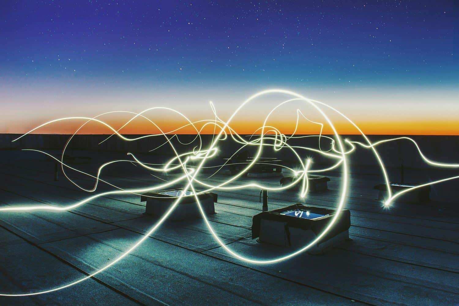 Long exposure photo of light trails on a rooftop at dusk, with a clear sky transitioning from blue to orange on the horizon.