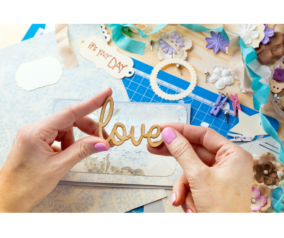 Hands holding a wooden "love" cutout surrounded by crafting materials, including paper, ribbon, and decorative flowers.