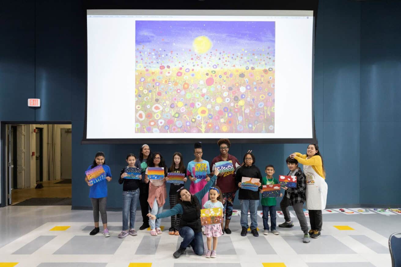 A group of children and adults stand in a room holding colorful paintings. A large artwork of a vibrant flower field is displayed on a screen behind them.