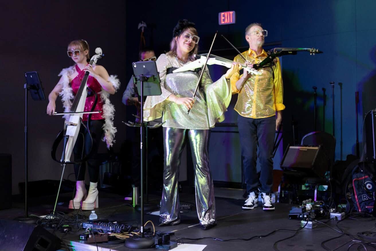 A trio of musicians performs on stage, featuring a cellist, a violinist, and an electric violinist. They are wearing vibrant outfits and are surrounded by musical equipment.