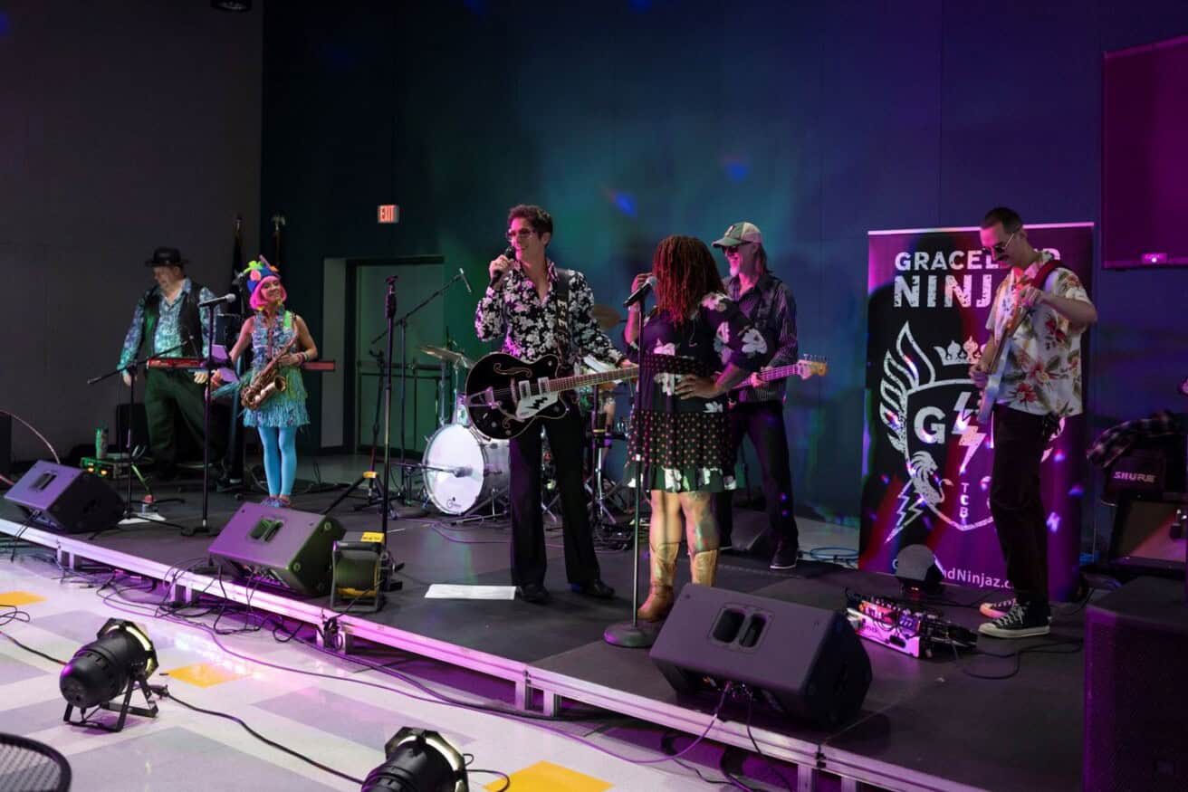 A band performs on stage with various instruments. Members wear colorful outfits. A backdrop displays "GRACELAND NINJA." Stage lights create a lively atmosphere.