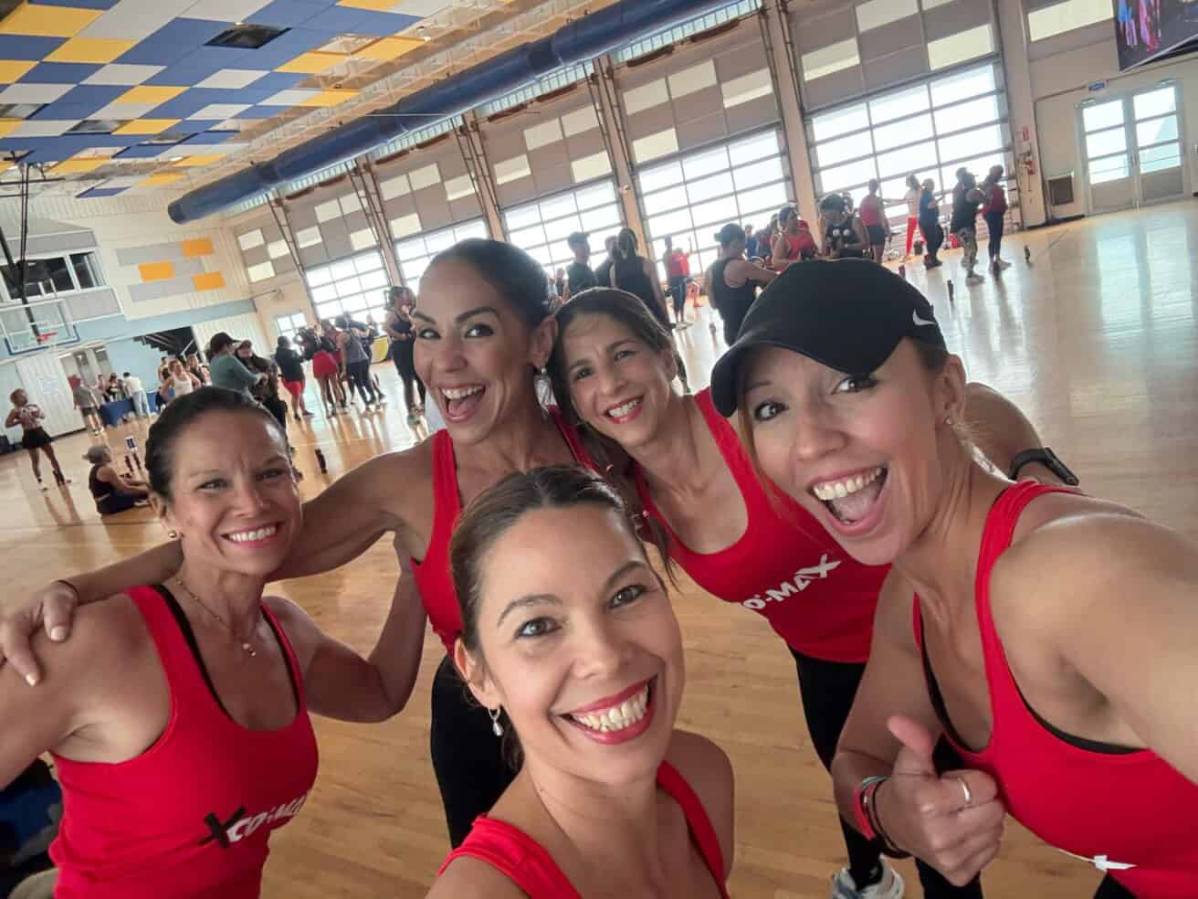 Group of five smiling women in red tank tops posing indoors during a fitness event with other participants in the background.