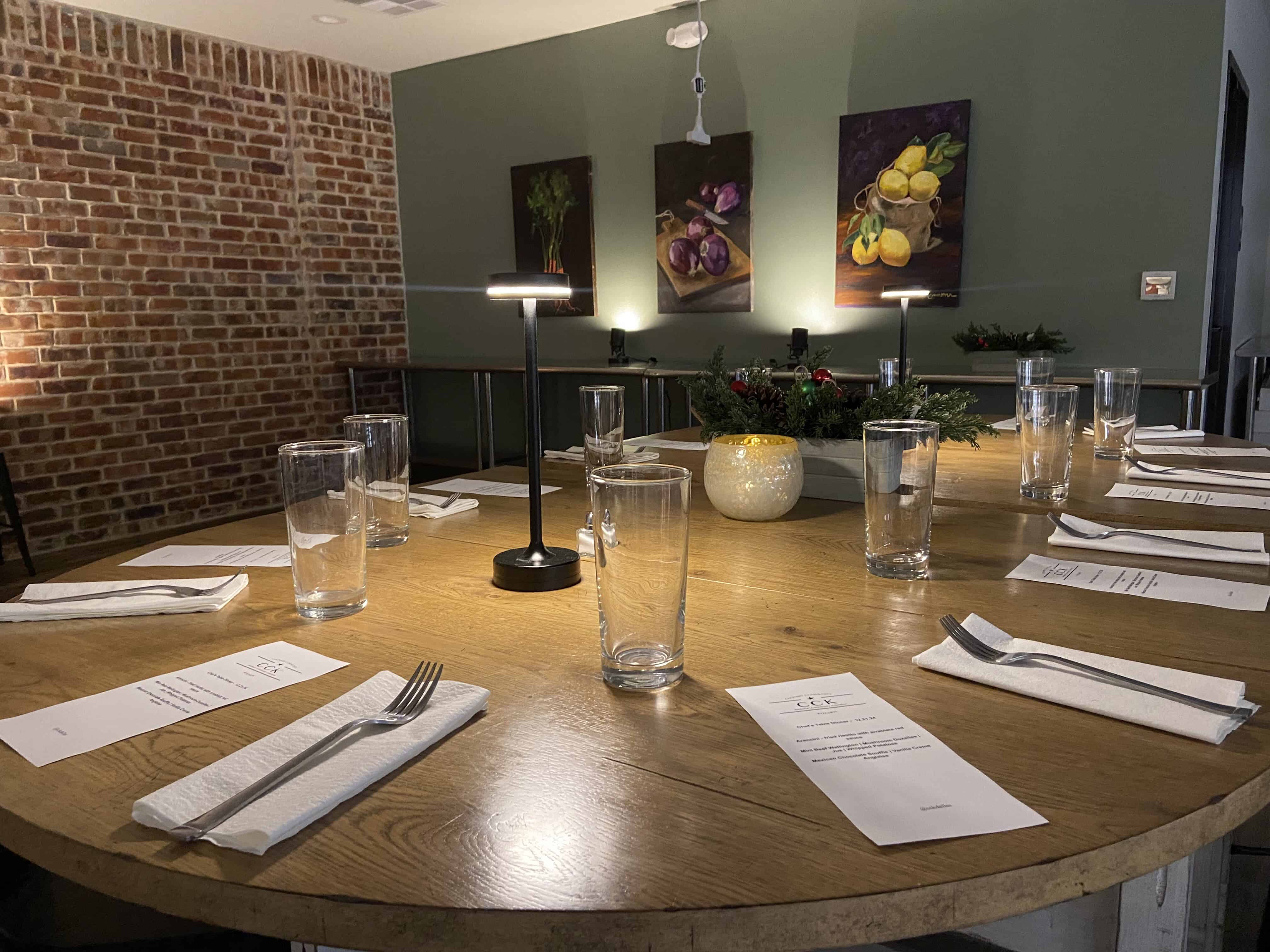 A round wooden table set for six with glasses, menus, and napkins, lit by small lamps. Brick wall and fruit paintings in the background.